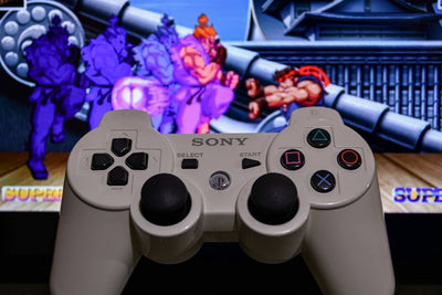 White Sony PlayStation controller in the foreground, with a TV screen behind showing a retro fighting game scene featuring characters mid-combat.